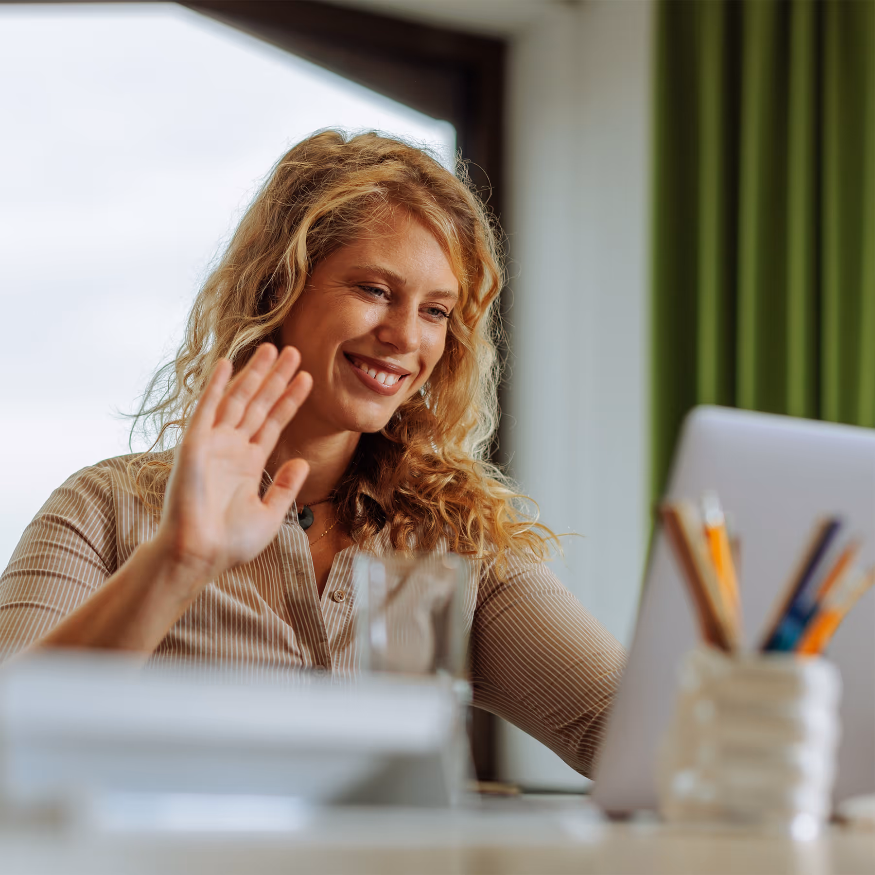 Smiling woman with curly blonde hair waving at a tablet during a video call indoors.