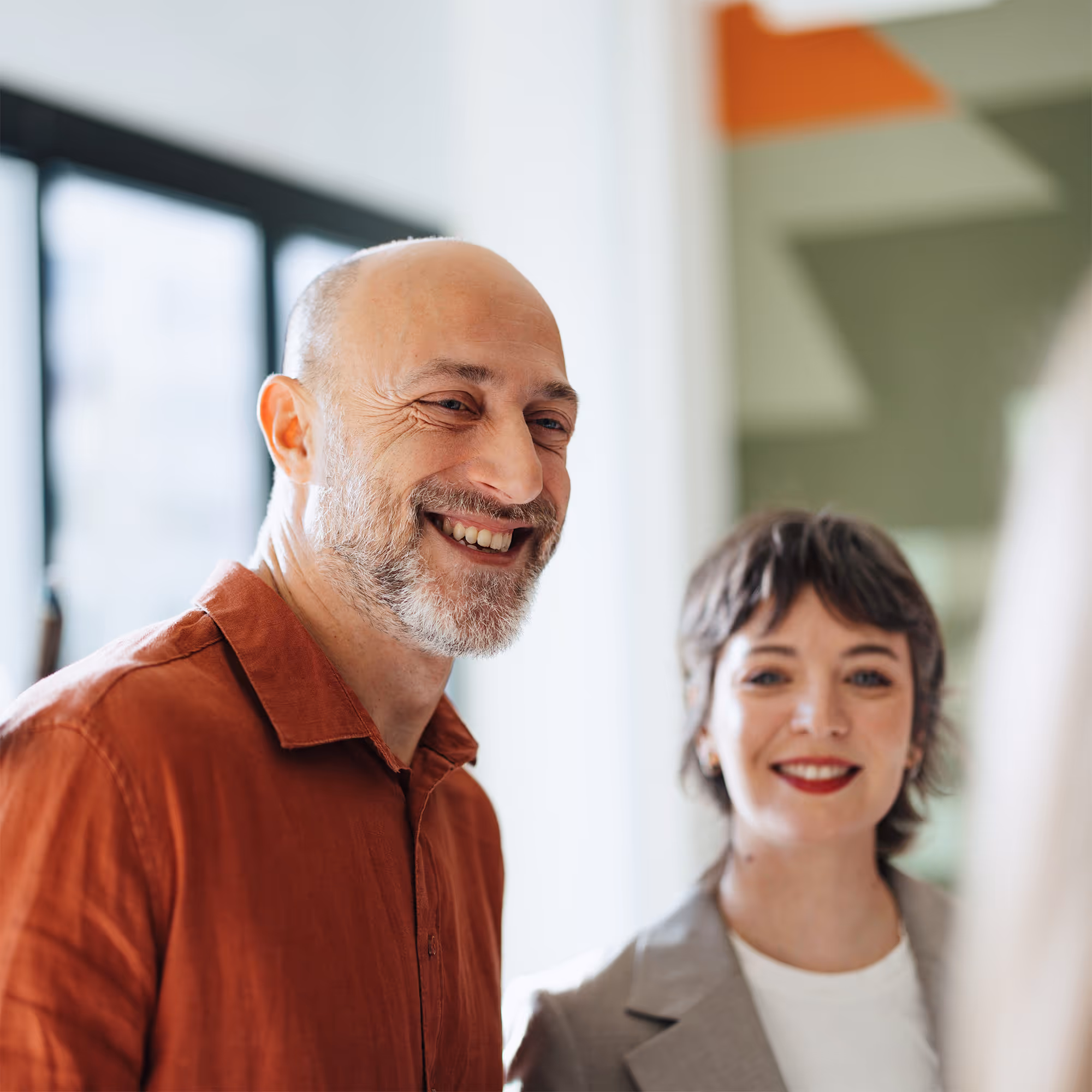 Smiling middle-aged man with a beard wearing a rust-colored shirt and a smiling woman with short hair and red lipstick in a gray blazer.
