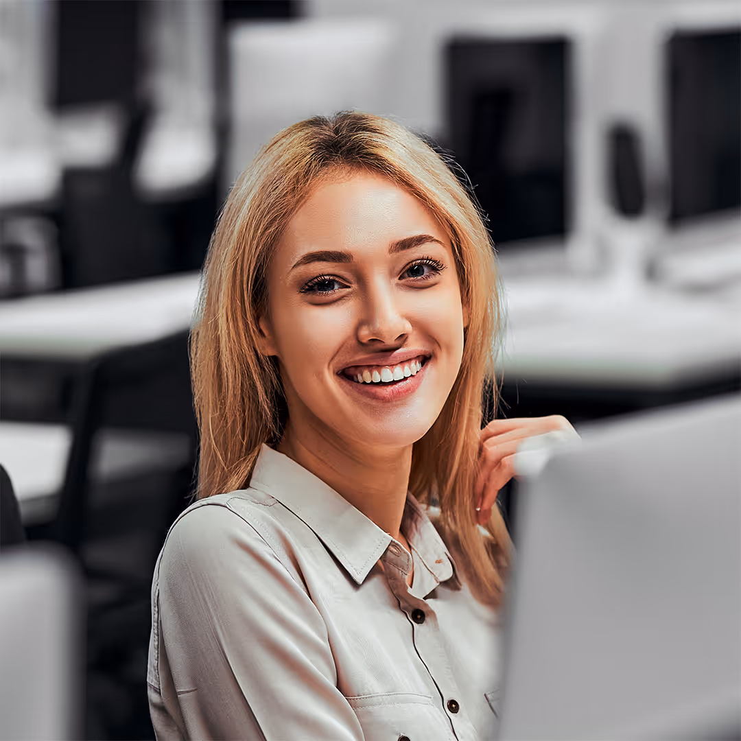 Smiling young woman with blonde hair sitting in an office environment with a laptop.