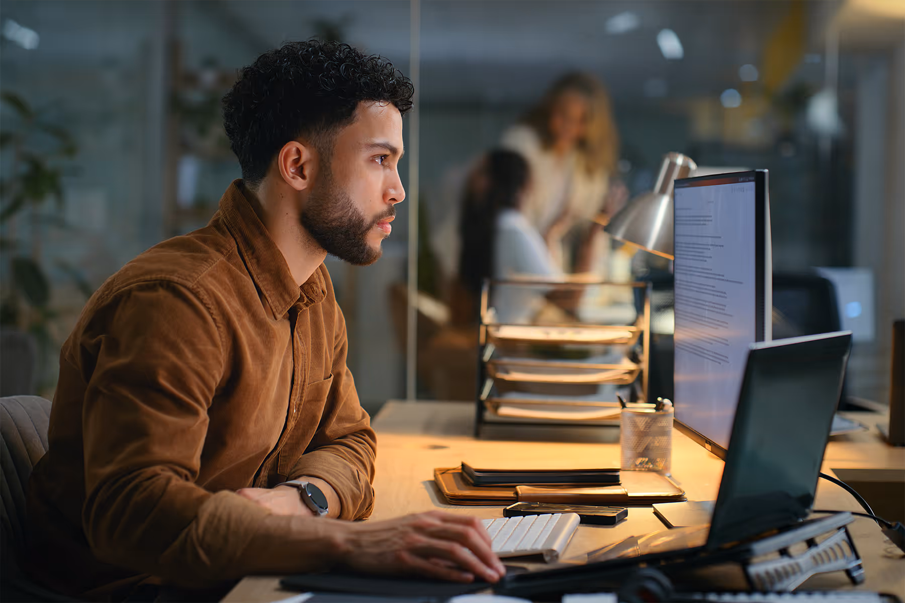 Man in a brown shirt working at a desk focused on a computer screen in an office with blurred people in the background.