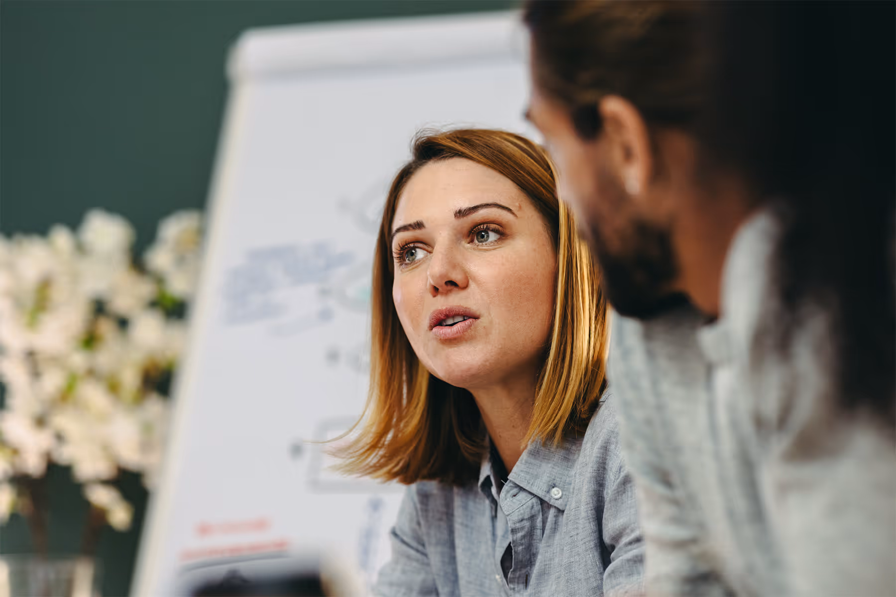 Close-up of a woman with shoulder-length hair talking to a man with long hair in an office setting with a whiteboard in the background.