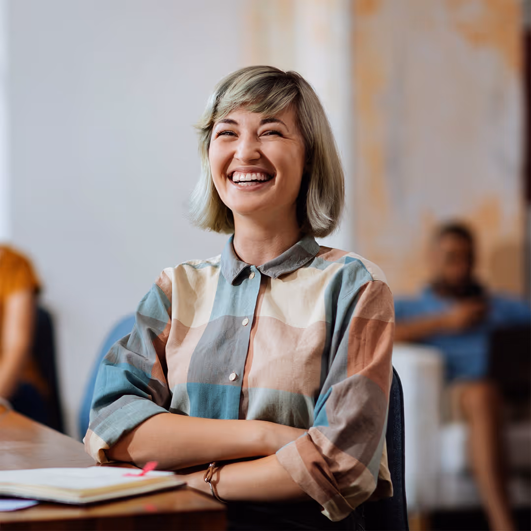 Smiling woman with short blonde hair wearing a multicolored checkered shirt, sitting at a desk with arms crossed.