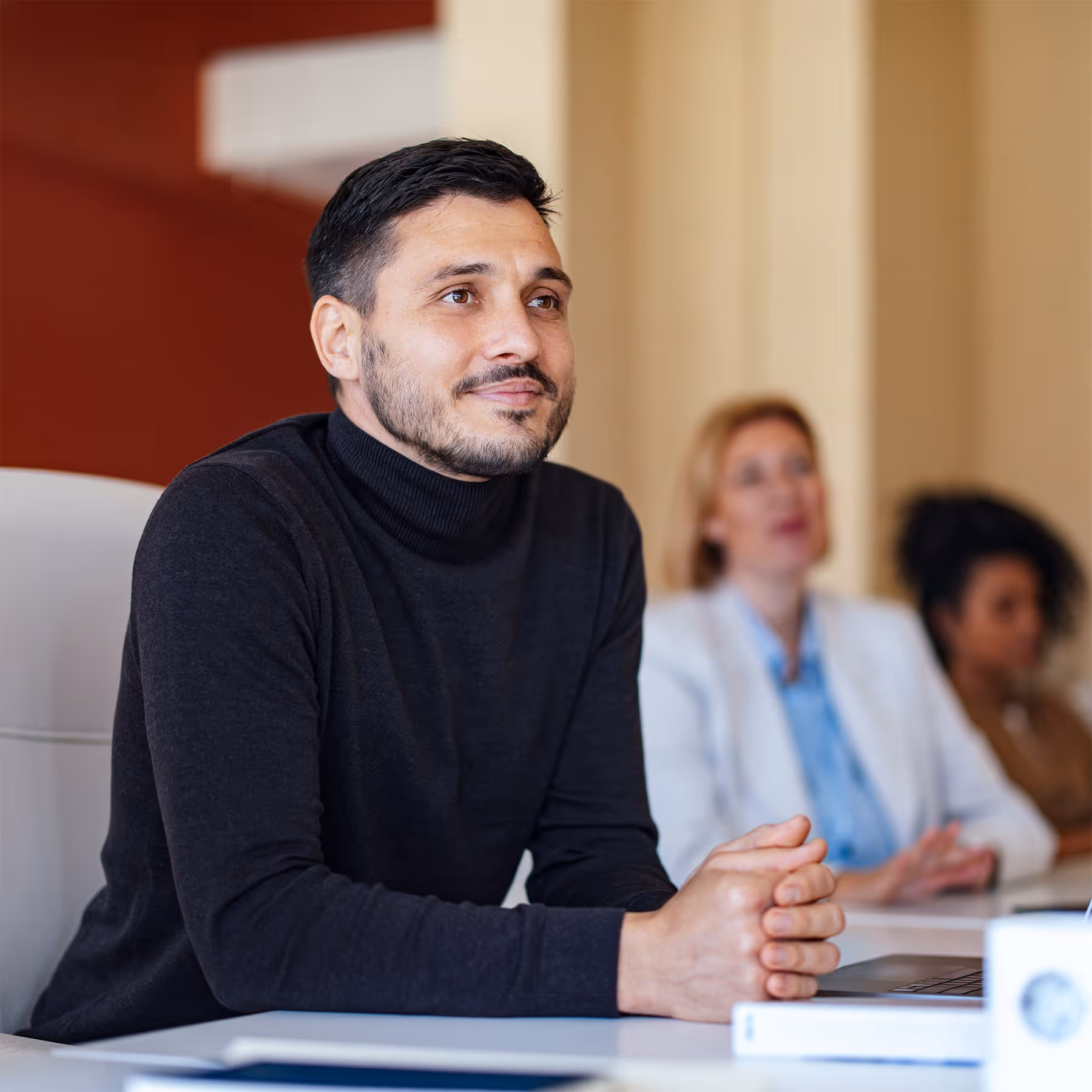 Man in black turtleneck sitting at a desk with two blurred women in the background.