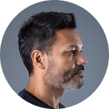 Profile portrait of a man with black hair and a salt-and-pepper beard against a gray background.