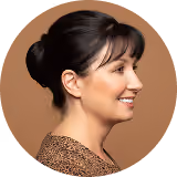 Profile of a woman with dark hair in a bun, smiling and wearing a brown patterned top.