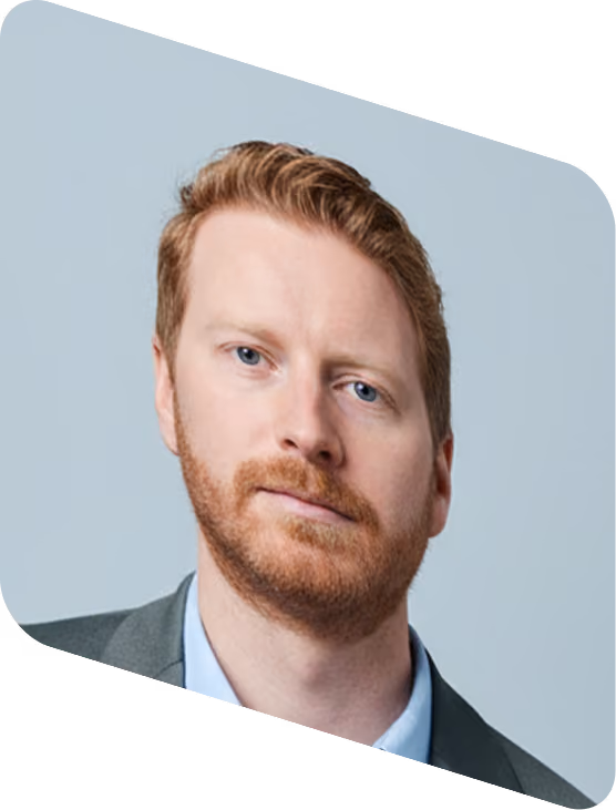 Portrait of a serious red-haired man with beard in a suit jacket and light blue shirt.
