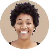 Smiling woman with short curly hair wearing a gray top against a beige background.