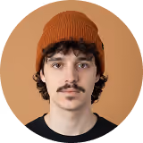 Young man with curly hair, mustache, wearing a brown beanie and black shirt on a tan background.
