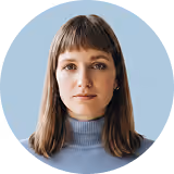 Portrait of a woman with brown hair and bangs wearing a blue turtleneck sweater against a blue background.