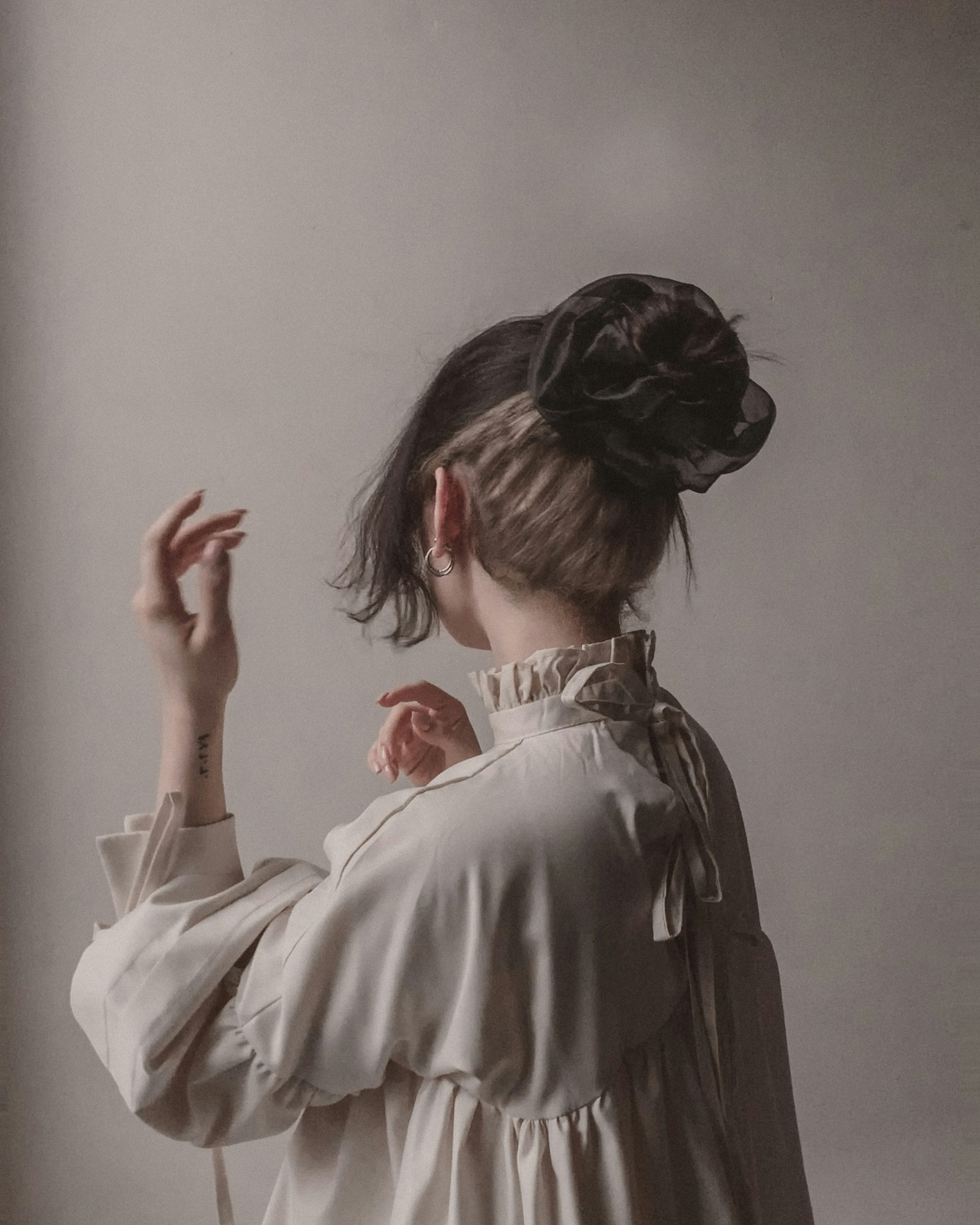 Woman seen from behind in a cream ruffled blouse, hair tied with a dark scrunchie, hands raised