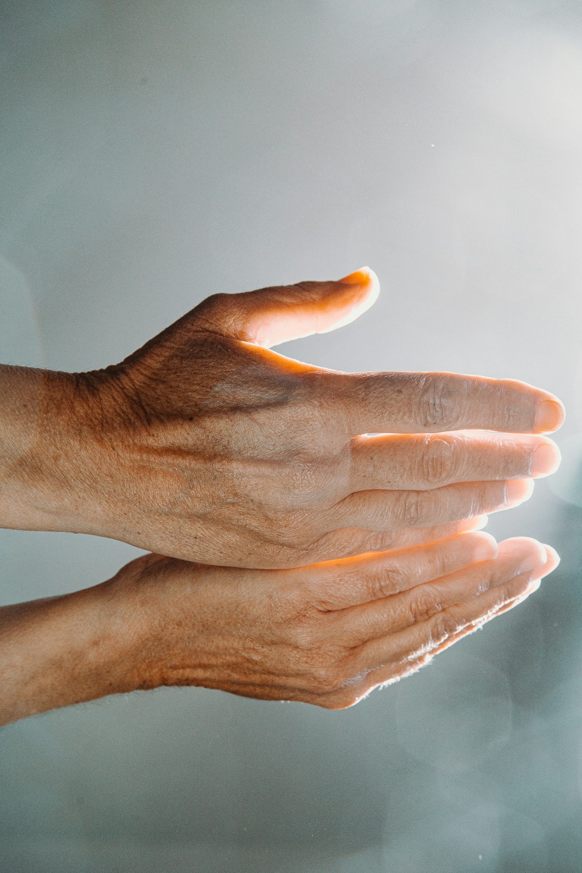 Two hands held together in warm backlight against a pale grey-blue background
