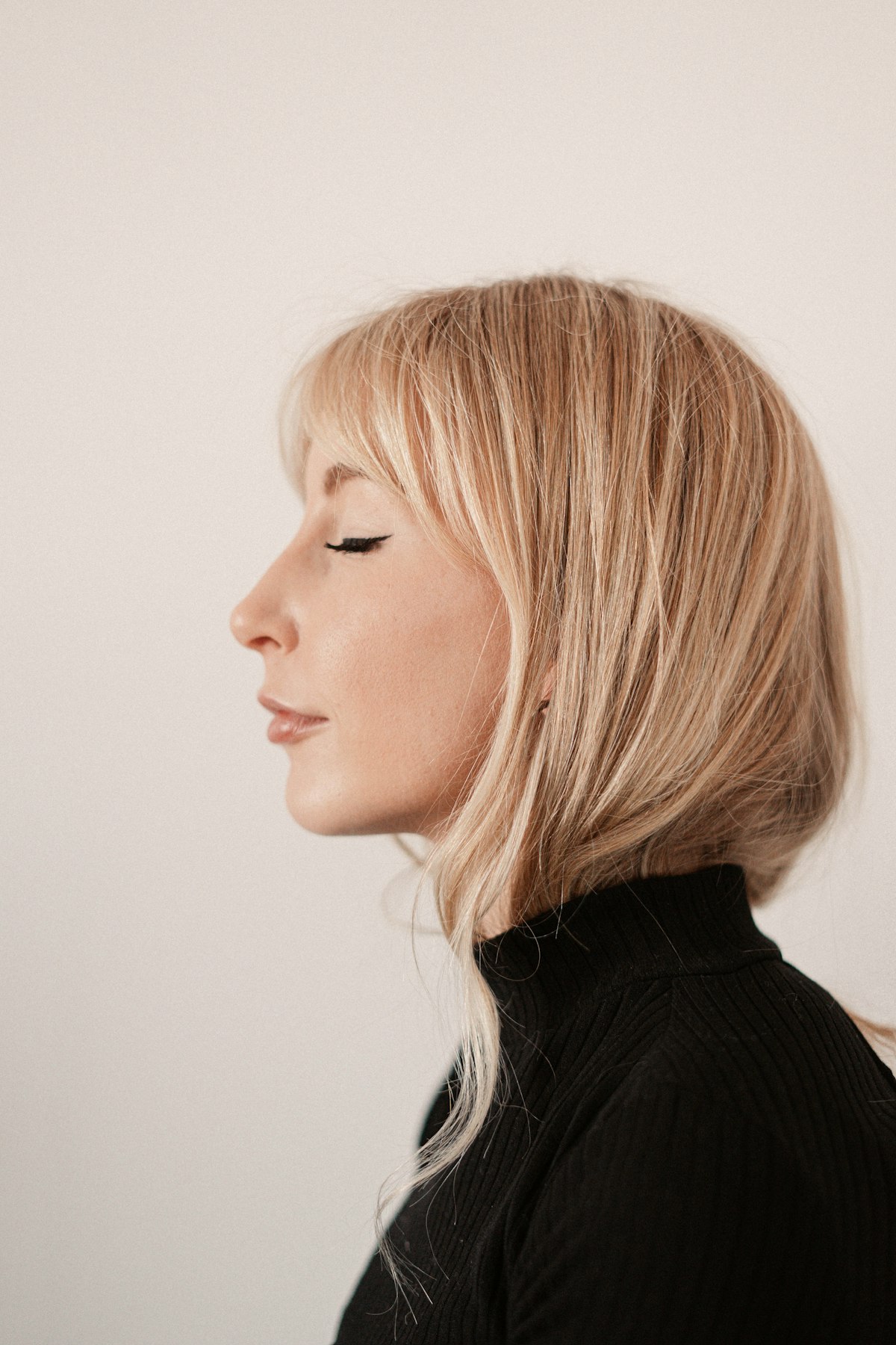 Profile portrait of a blonde woman with her eyes closed, wearing a black turtleneck against a pale background