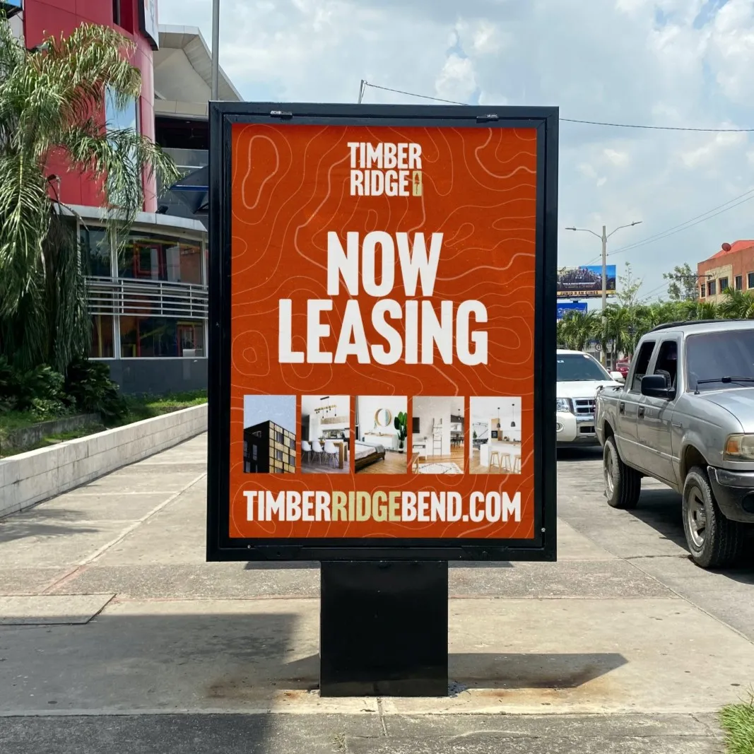 Outdoor advertisement board on a sidewalk showing Timber Ridge leasing information with photos of modern apartment interiors and exterior, and website timberridgebend.com, on a red background with contour lines.