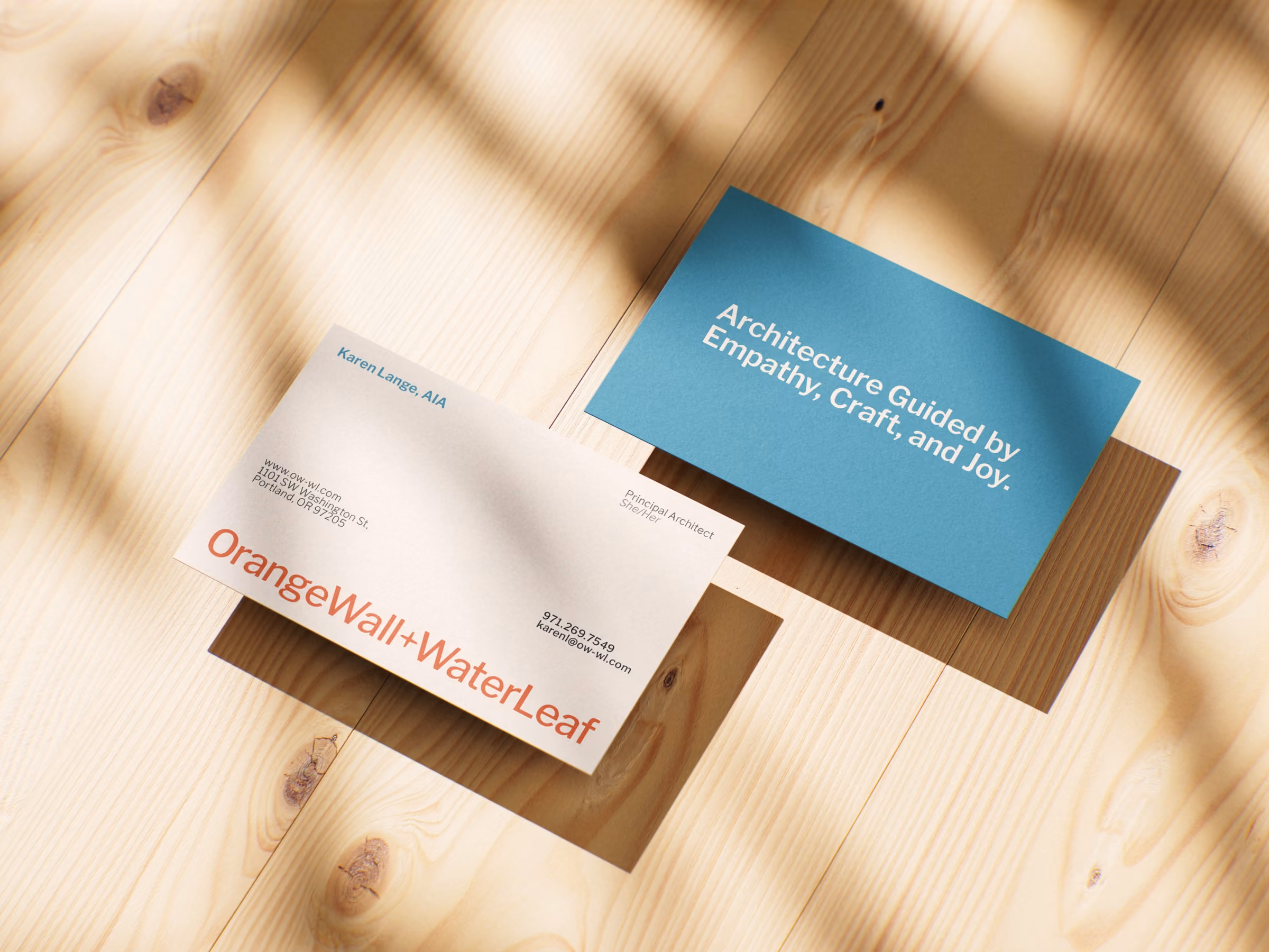 Two OrangeWall+WaterLeaf business cards on light wood surface with shadows.