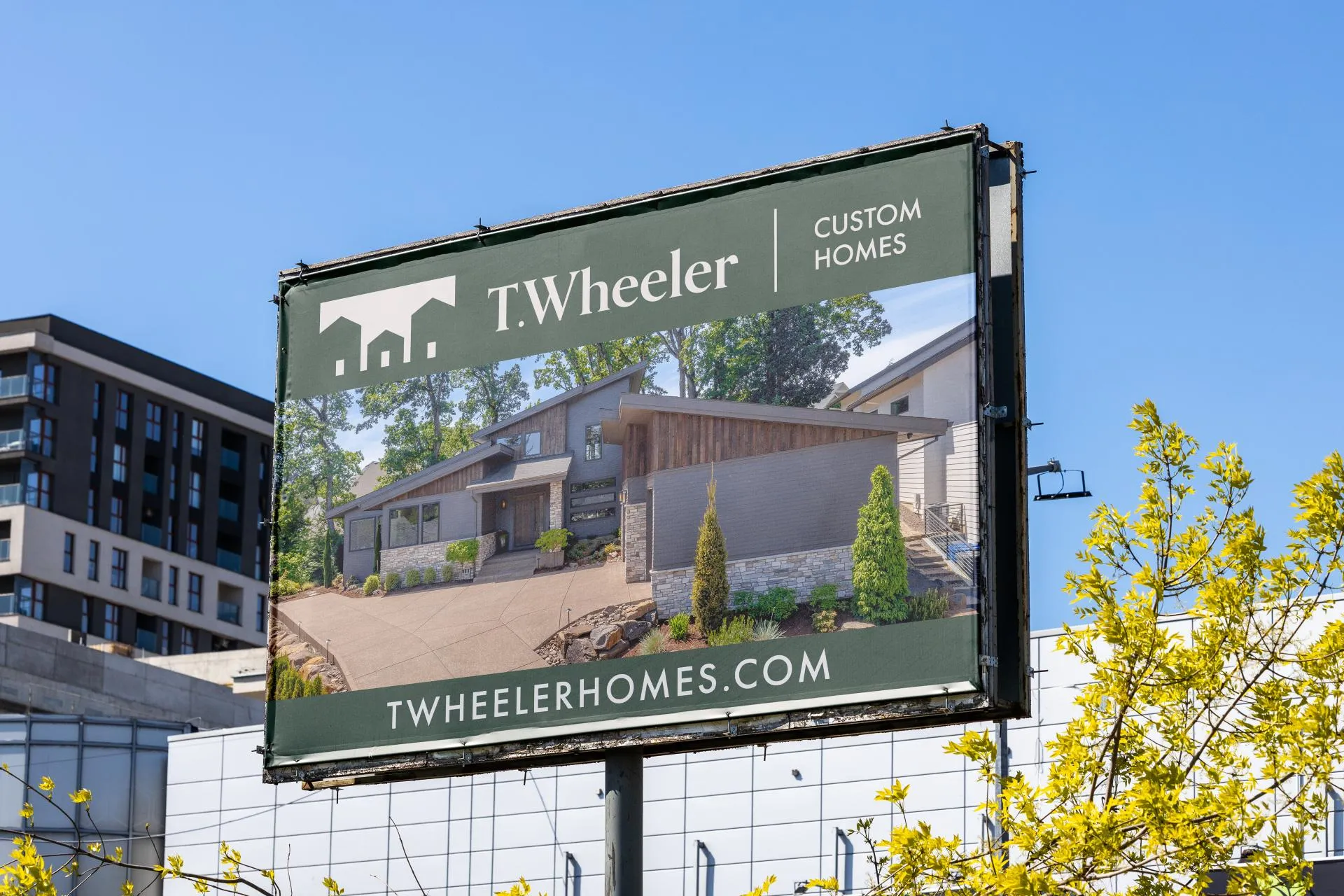 Outdoor billboard advertising T. Wheeler Custom Homes featuring a modern house with wood and stone exterior surrounded by trees, with the website TWHEELERHOMES.COM displayed at the bottom.
