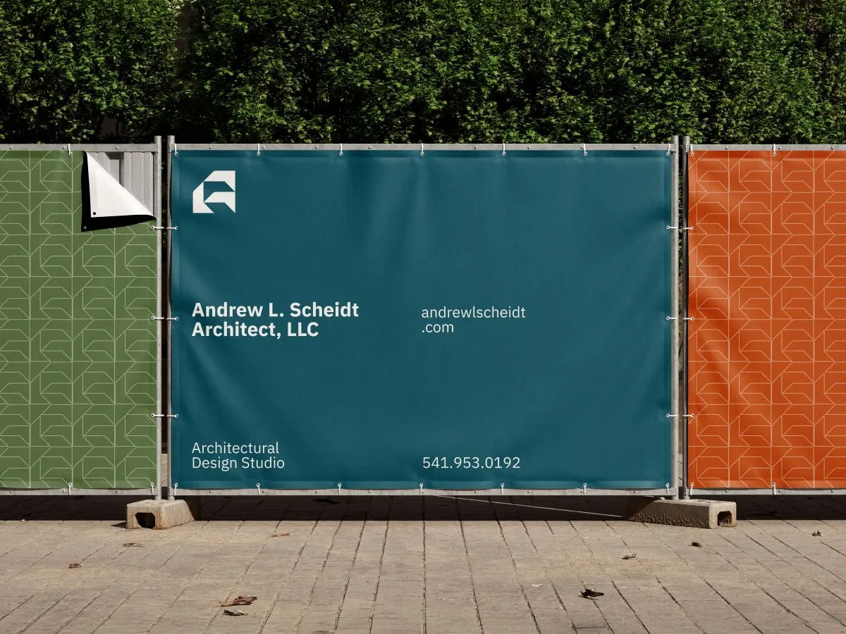 Construction site barrier with a teal banner displaying the text 'Andrew L. Scheidt Architect, LLC' along with the website 'andrewlscheidt.com,' the description 'Architectural Design Studio,' and the phone number 541.953.0192, flanked by green and orange patterned panels.