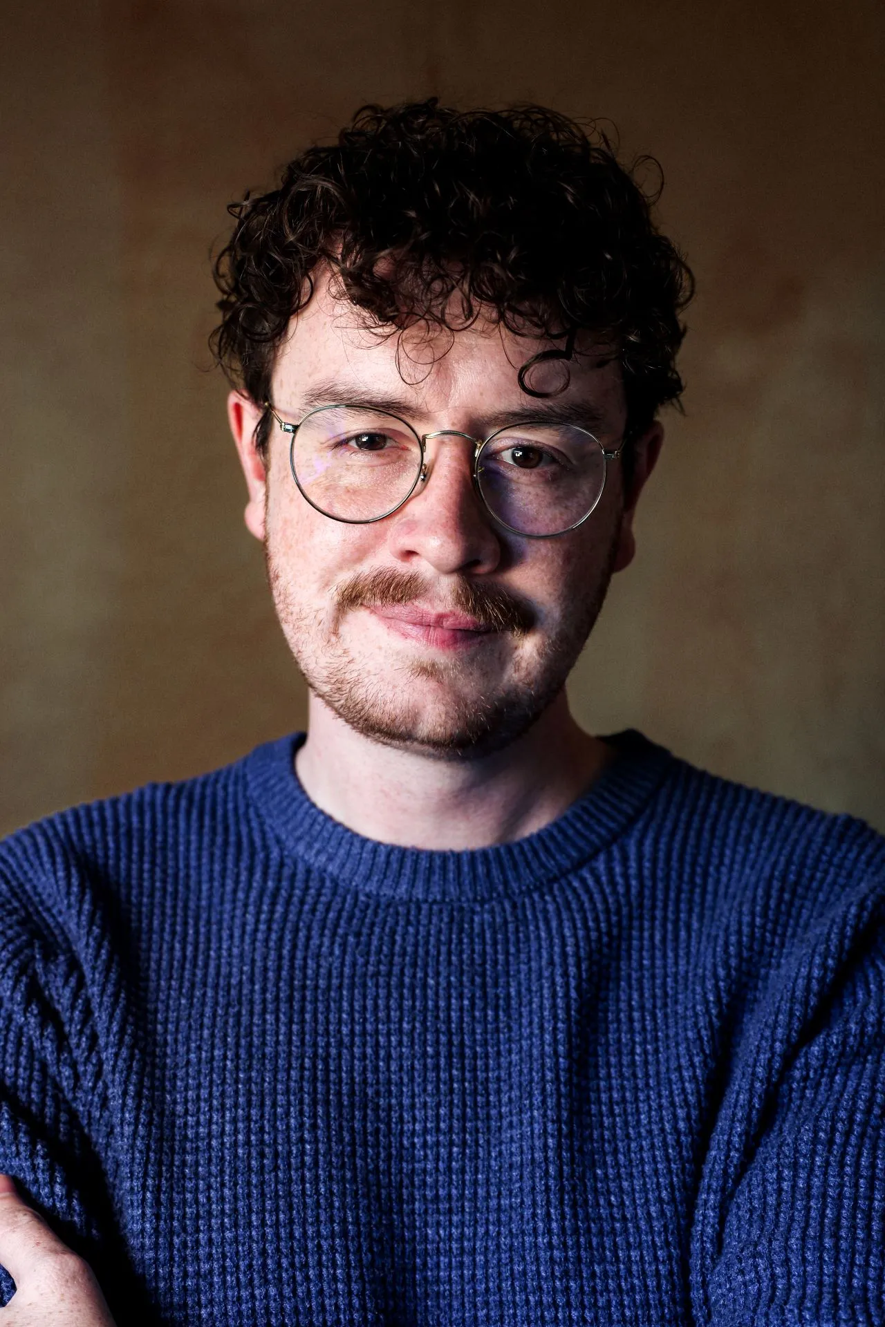 Smiling professional man with glasses wearing a sweater standing in front of a photo background.