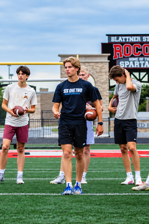 Young quarterbacks holding footballs during an outdoor training session at a football stadium.