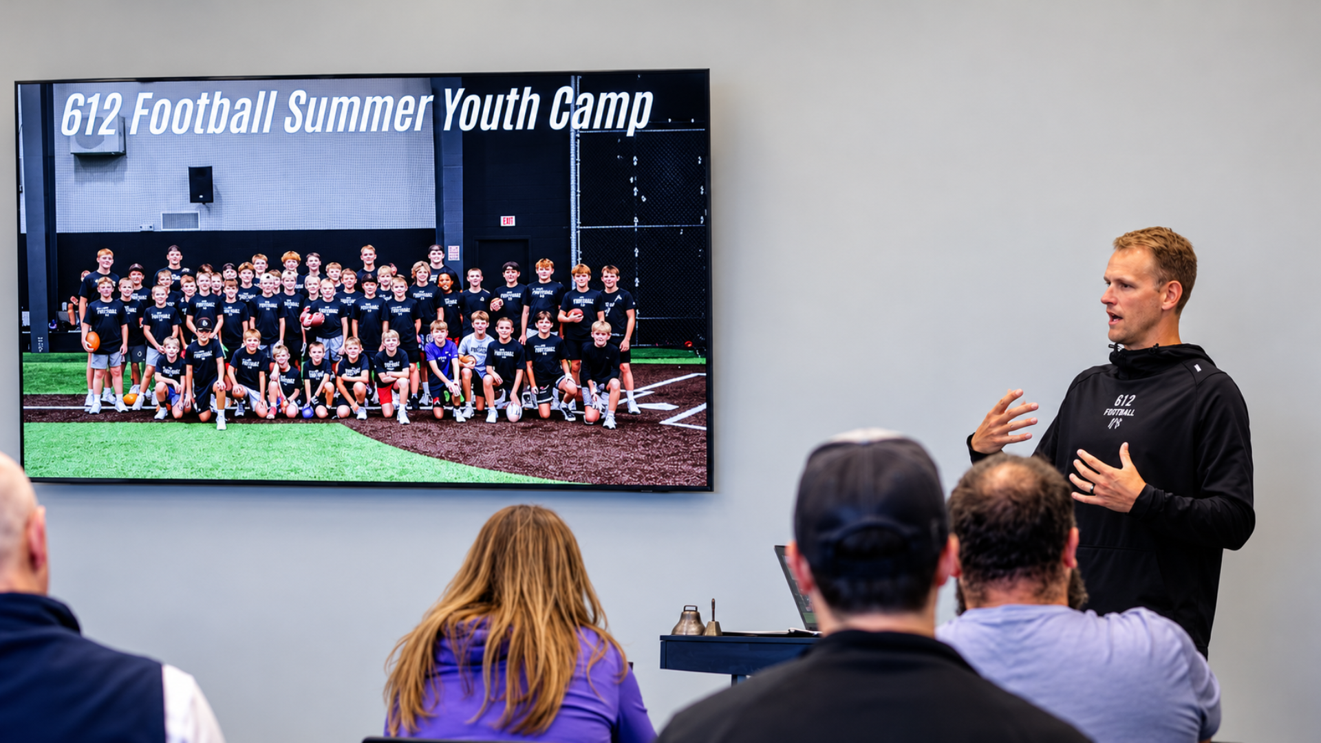 612 Football coach presenting to families at the Summer Youth Camp, focused on the coach rather than the background screen.