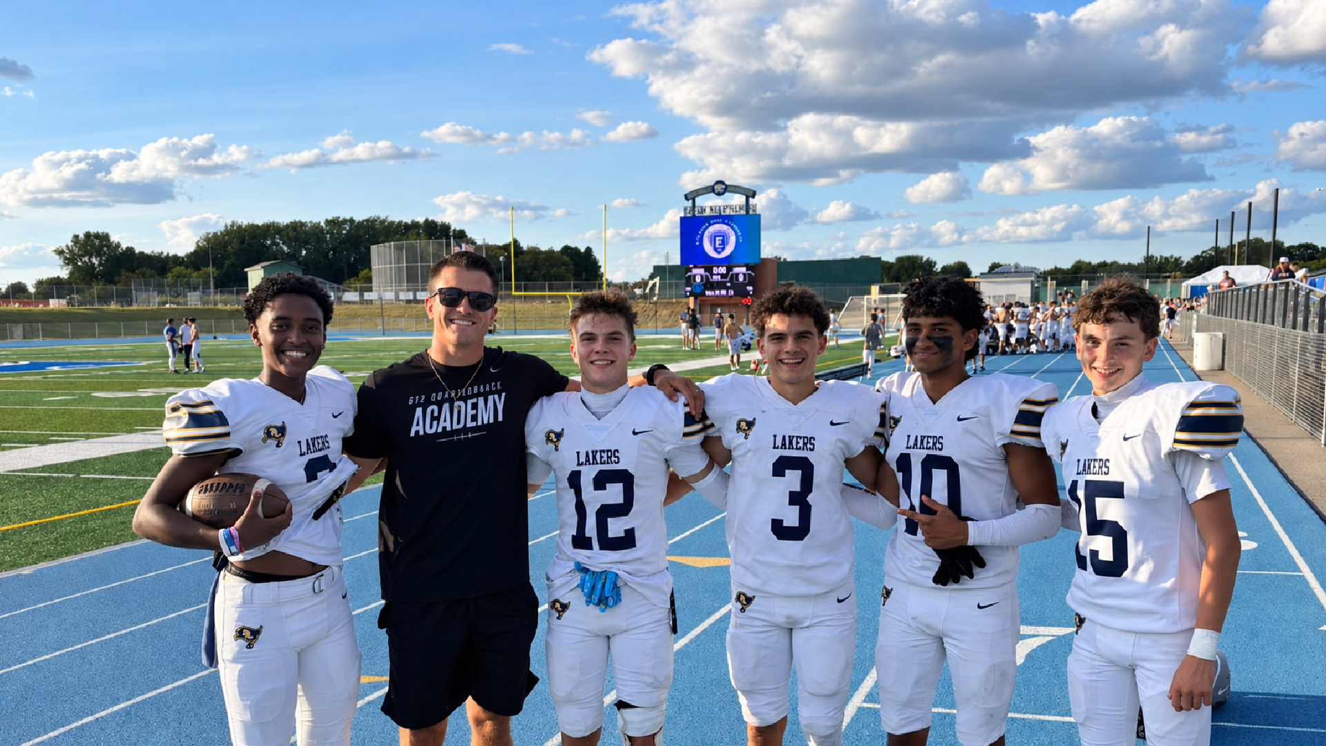 Football coach posing with five high school players in uniform on a football field after a game.