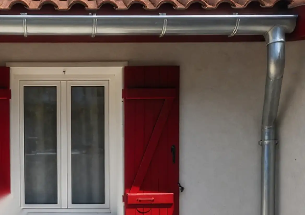 Gouttière zinc et sa descente au-dessus d'une fenêtre à volets rouges, secteur de Bayonne