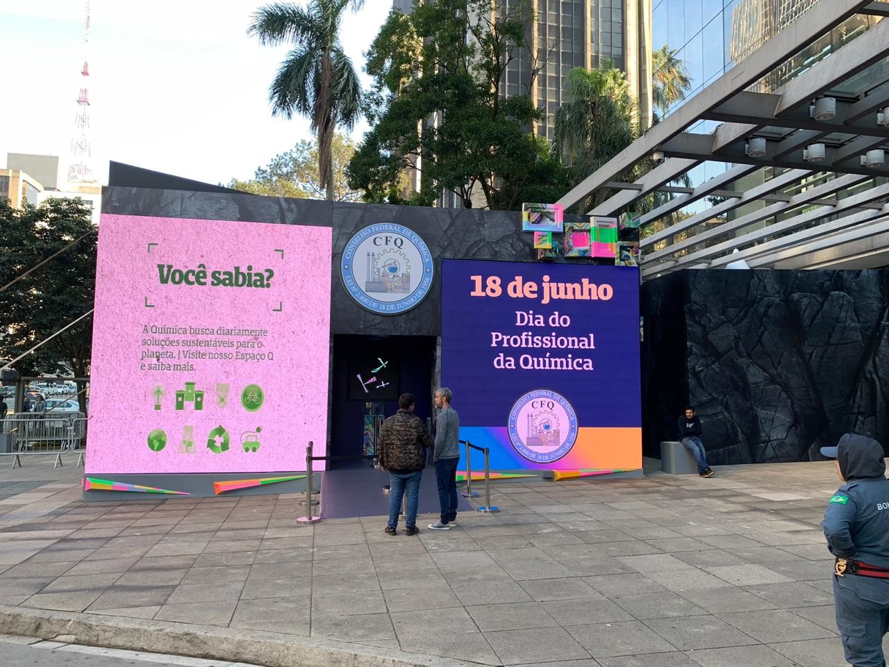Pink Você sabia? panels and the 18 June Chemist's Day wall on the side of the pavilion, a visitor reading