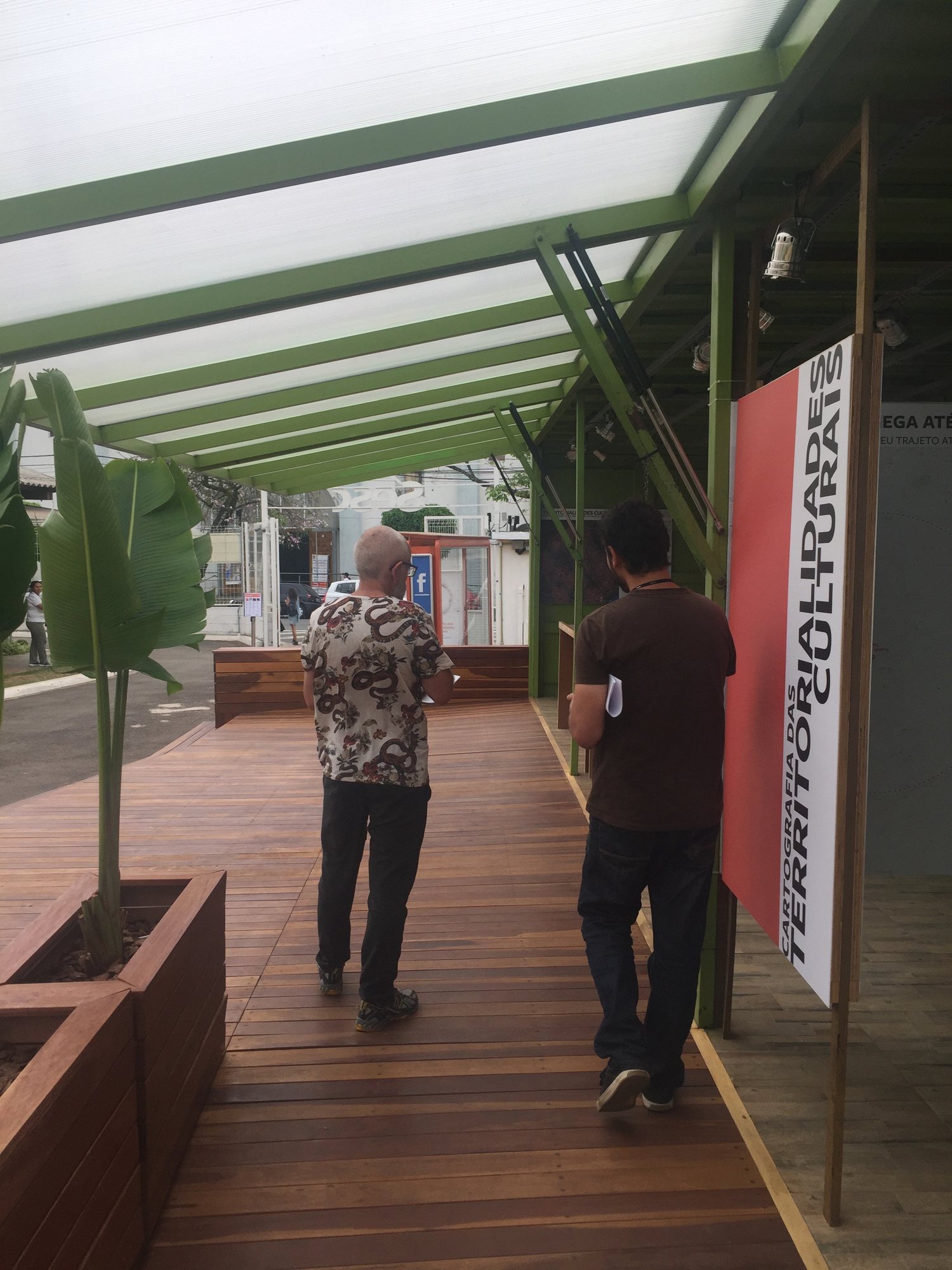 Sesc · 2017 · Sesc Verão — Two visitors walking under the timber pergola, between the planted deck and the pavilion wall.