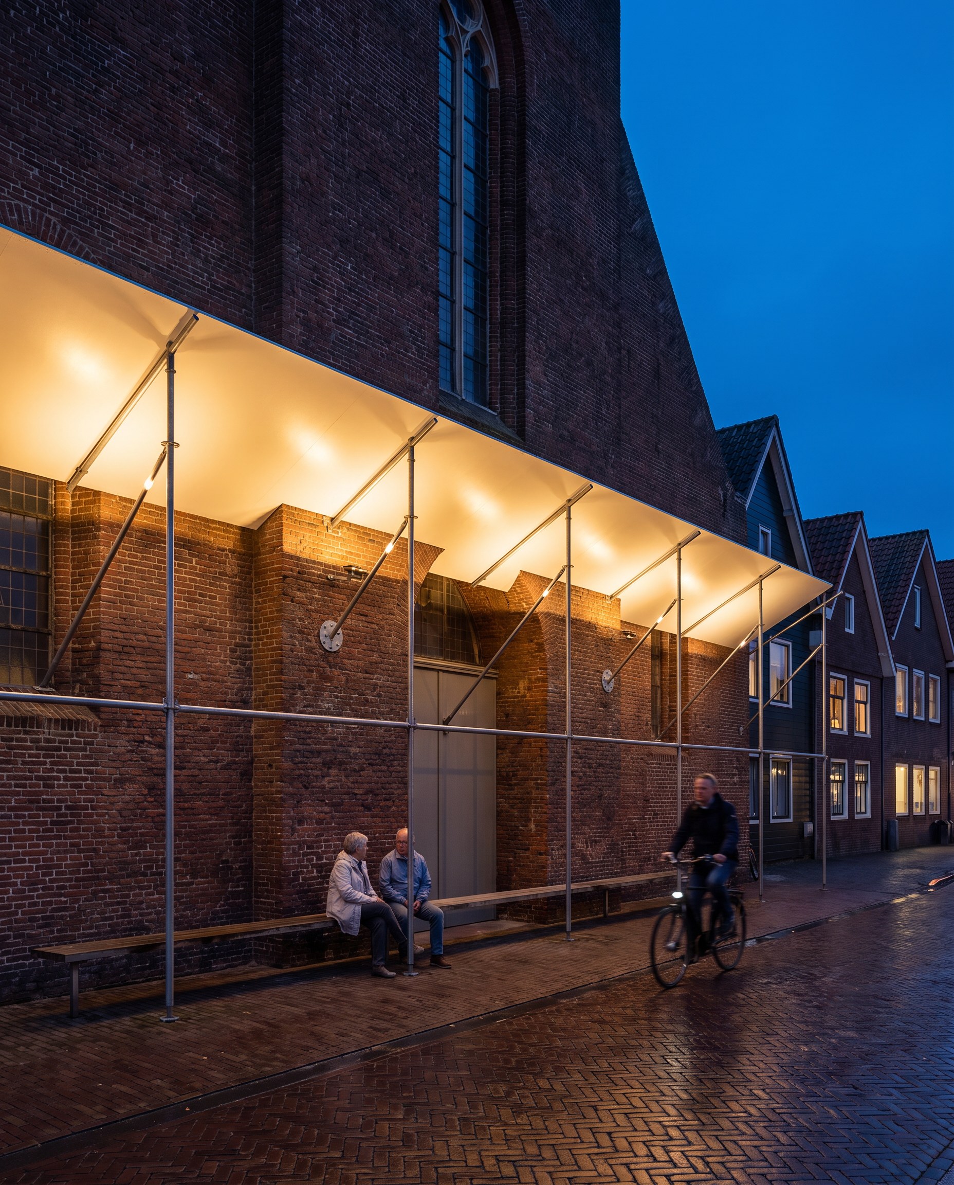 The catch-fan on the dike street lit as a porch, two neighbours on the long bench, a cyclist on the wet brick paving, the tower rising out of the frame