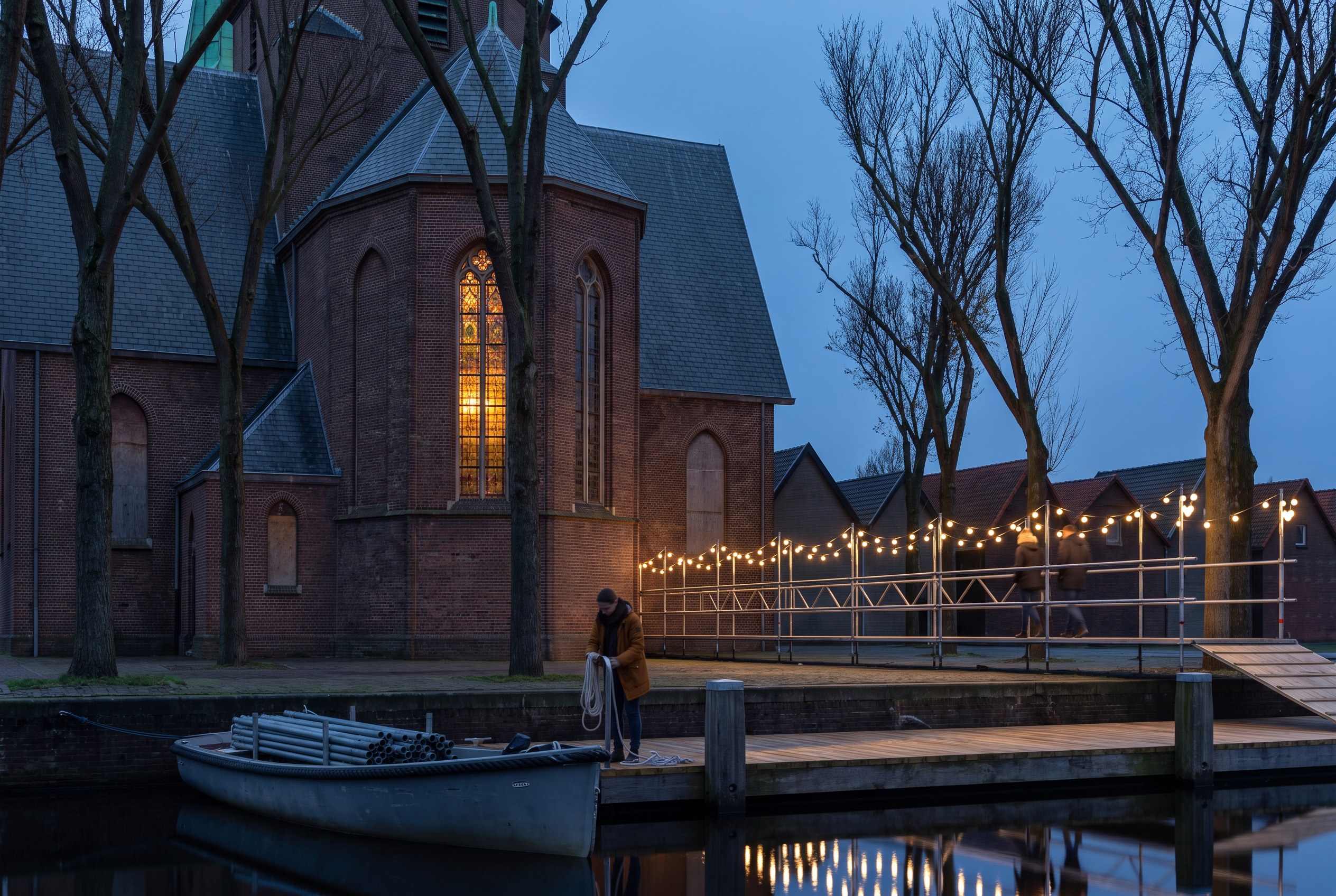 Blue hour on the Nieuwendammerham: a boat loaded with scaffold tubes at the floating pier, a walkway of warm bulbs leading to the dark brick choir of the church