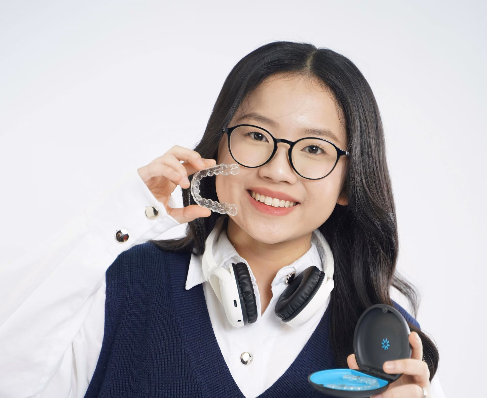Woman with glasses holding a clear dental aligner and an open case, wearing headphones and smiling against a white background