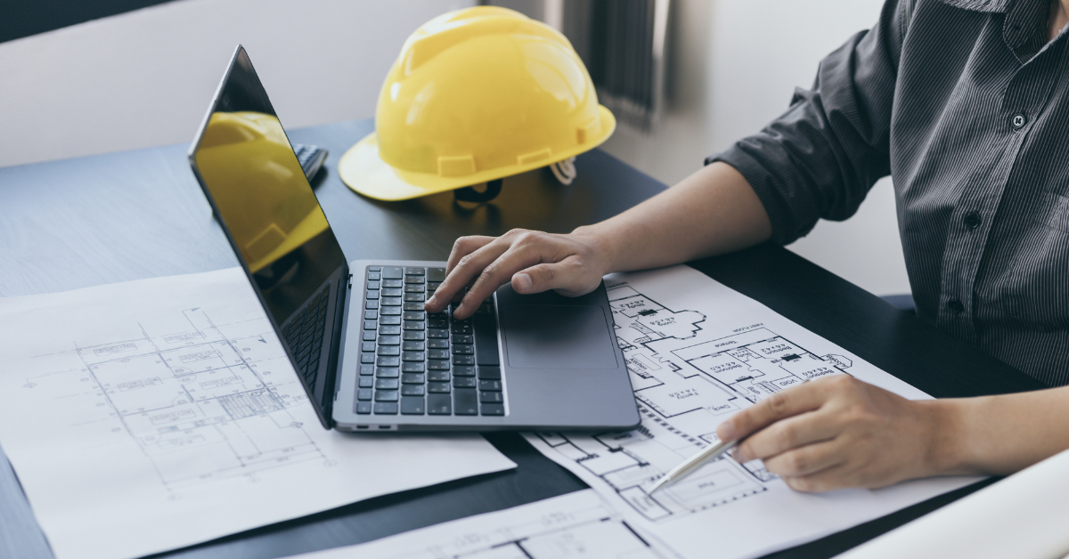 A laptop open on project plans beside a hard hat