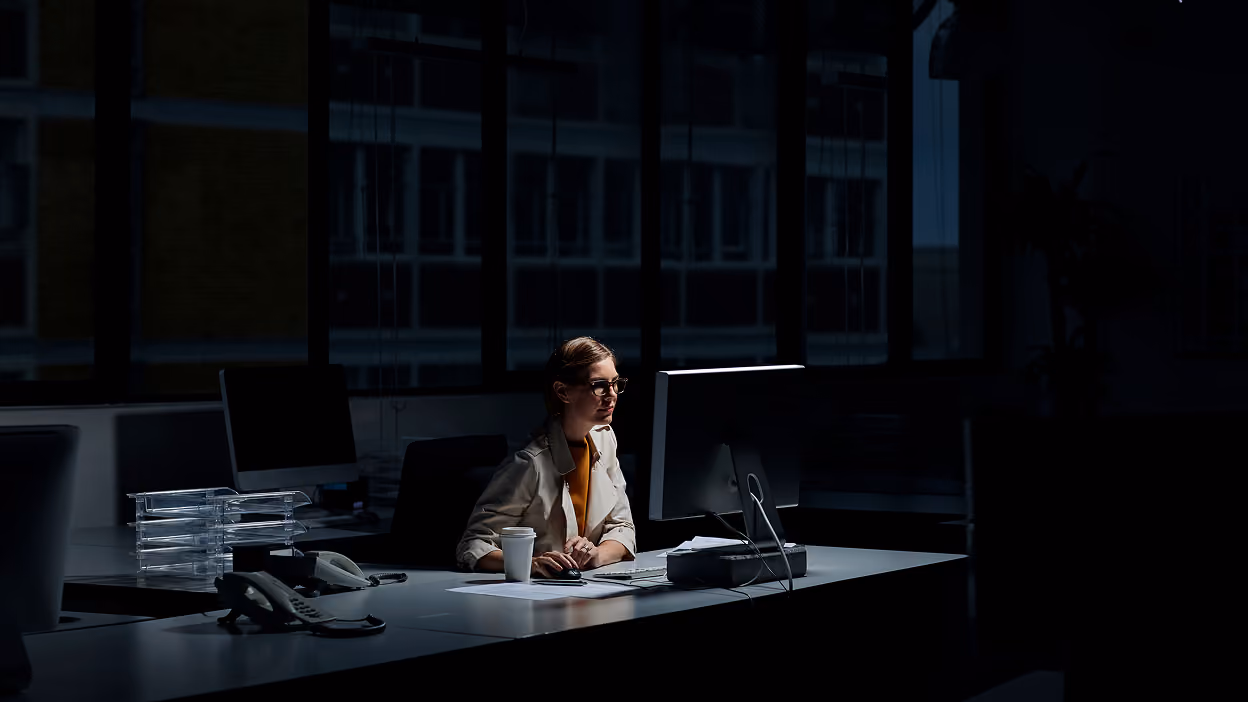 Woman with glasses sitting at a desk working on a desktop computer in a dimly lit office at night, with a coffee cup and office supplies on the desk.