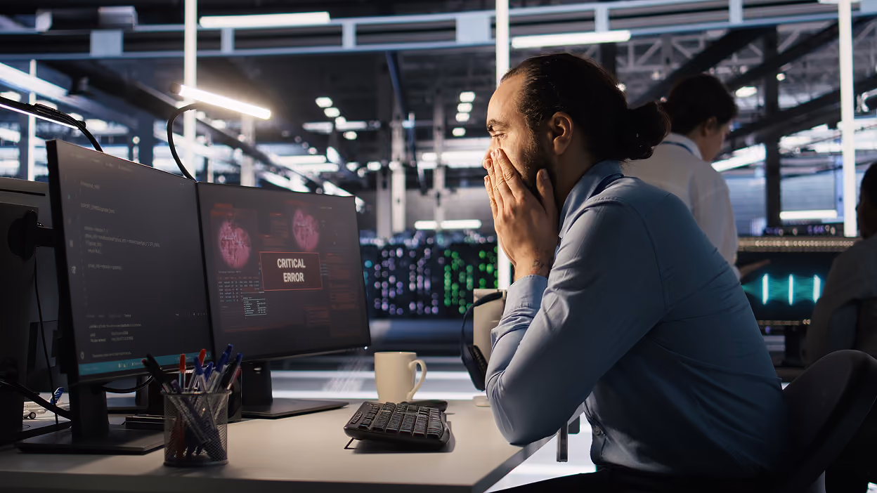 Man in a blue shirt sitting at a desk with dual monitors showing a critical error message, looking stressed and with hands covering his mouth in an office with others working in the background.