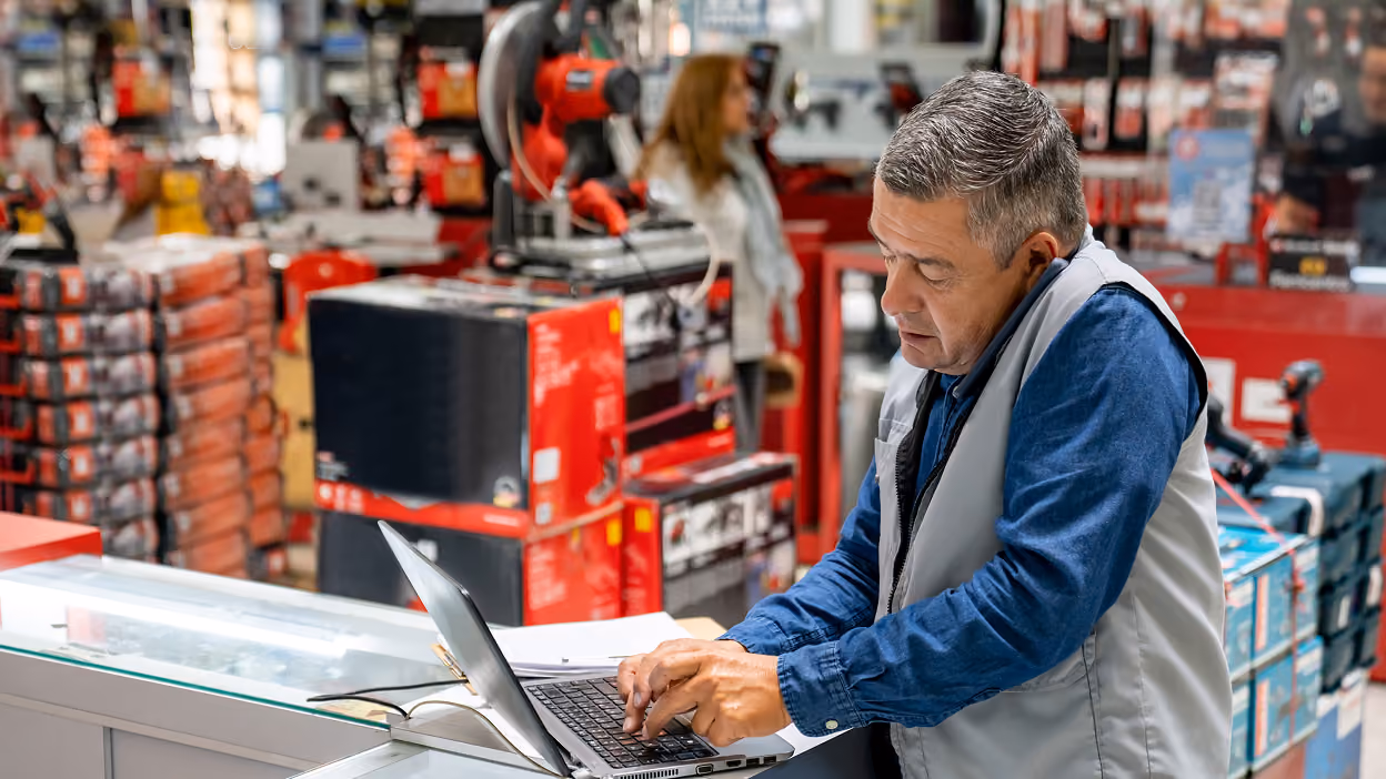 Middle-aged man in a gray vest and blue shirt working on a laptop at a counter in a hardware store with shelves of tools and equipment in the background.
