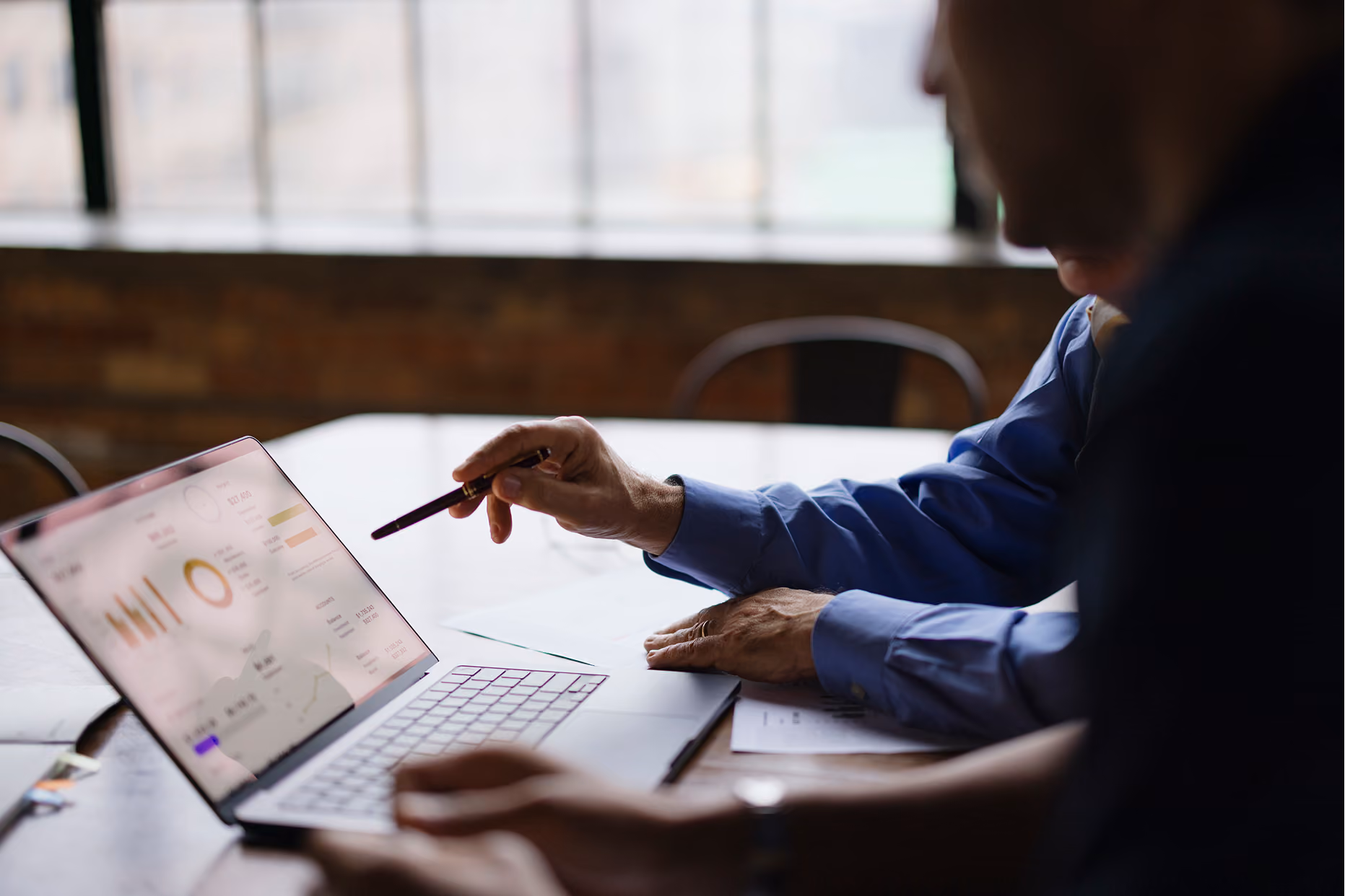 Two people sitting at a table looking at a laptop screen displaying graphs and charts; one person points at the screen with a pen.