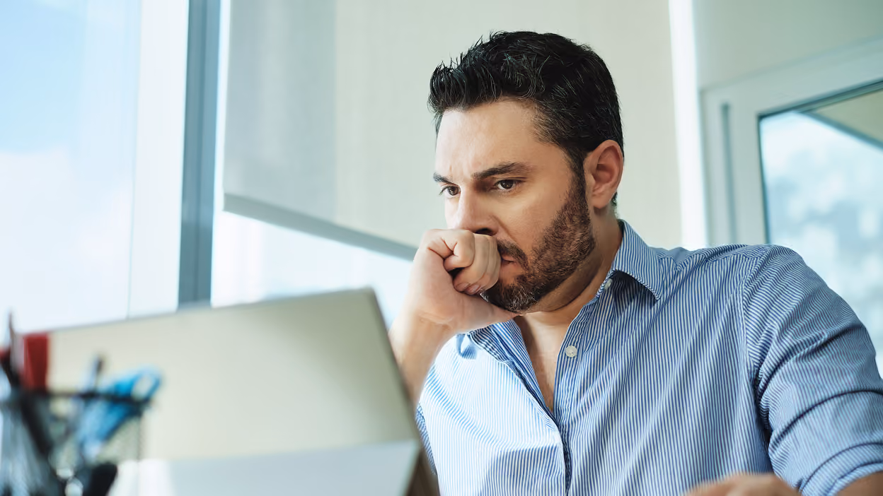 A serious man with dark hair and a trimmed beard wearing a blue striped shirt looks intently at a laptop screen indoors with large windows behind him.