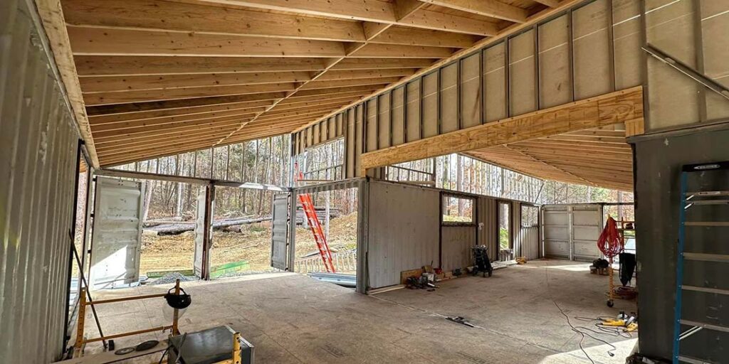Interior of a large unfinished building with exposed wooden beams and open shipping container doors.