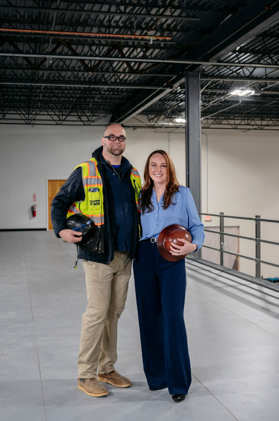 Man in safety vest and woman holding hard hats standing together in an industrial warehouse.