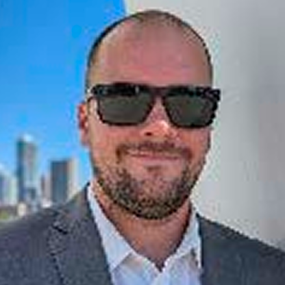 Man with a beard wearing black sunglasses, a suit jacket, and a white shirt outdoors with buildings in background.