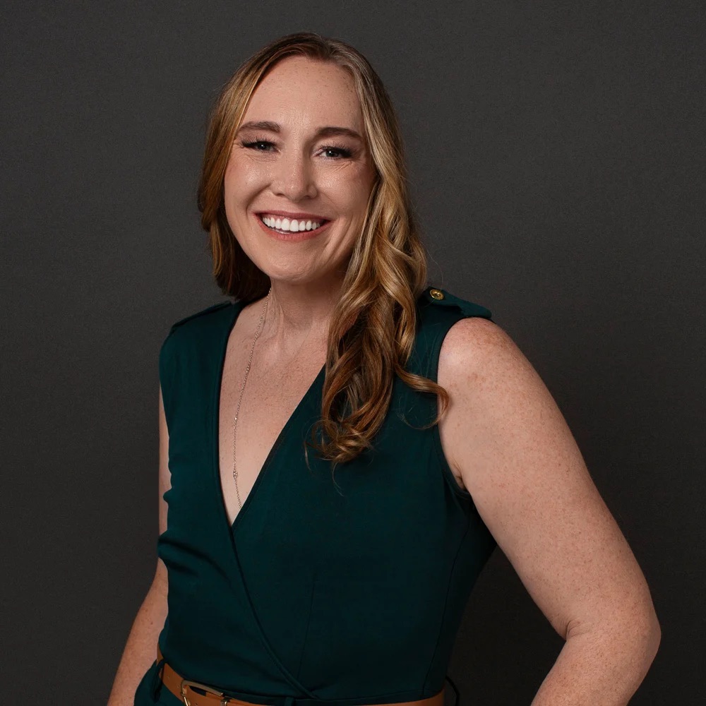 Smiling woman with long wavy hair wearing a sleeveless green dress and brown belt against a dark background.