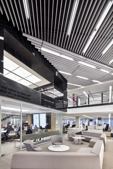 Modern office lounge area with gray sofas, round tables, glass-walled meeting rooms, and ceiling lights.