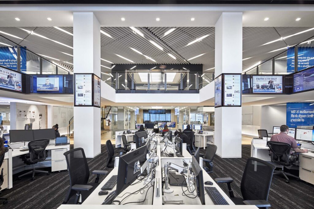 Modern open-plan office with multiple desks, computers, office chairs, and large wall-mounted screens.