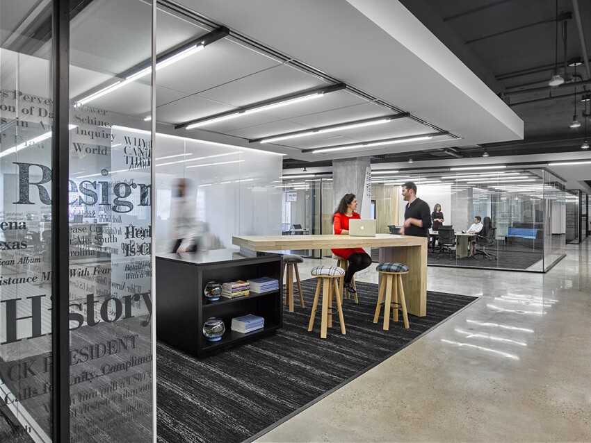 Modern office space with glass walls, people working and talking around a wooden table with stools.