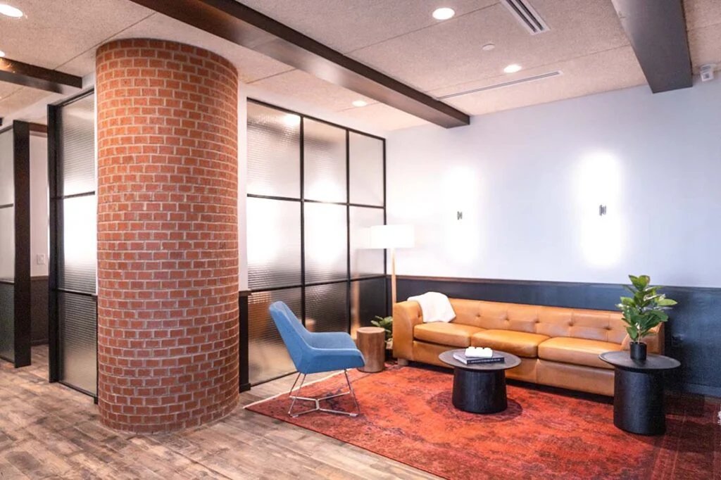 Modern lounge with brown leather sofa, blue chair, round tables, red rug, and brick column.