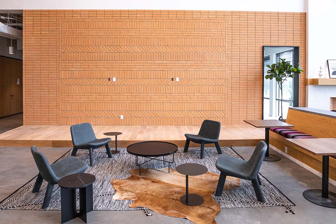 Modern seating area with four blue chairs around round black tables on rugs in front of a brick accent wall.