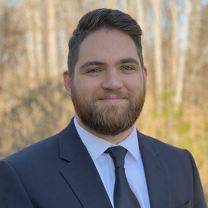 Man with beard wearing a dark suit and tie, smiling with a blurred outdoor background of trees.