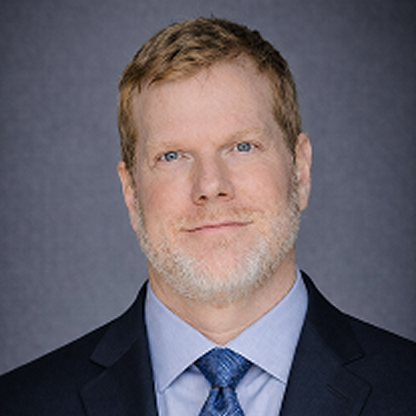Portrait of a man with short light brown hair and beard wearing a suit and blue tie.