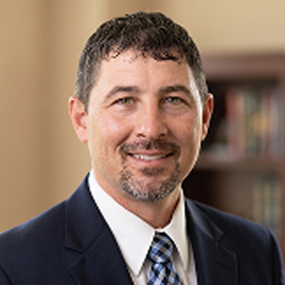 Smiling man with short dark hair and goatee wearing a suit and patterned tie in an office.