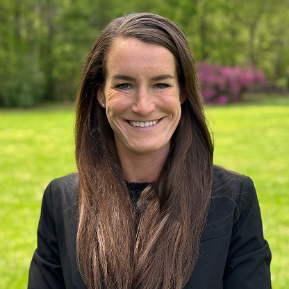 Smiling woman with long brown hair wearing a black blazer standing outdoors with green grass and trees behind her.