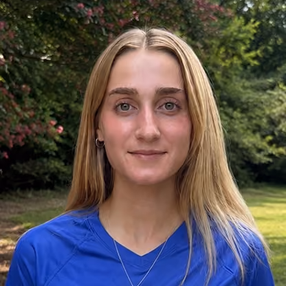 Young woman with long blonde hair wearing a blue shirt outdoors with greenery in background.